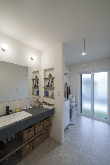 洗面の鏡も幅いっぱいのサイズ（1270mm）で設計しているので、夫妻が並んで使っても狭さを感じさせない。奥は室内干しスペースとなっていいて、天気の良い日は、そのまま右手のドアから外に出て外干しもできる。